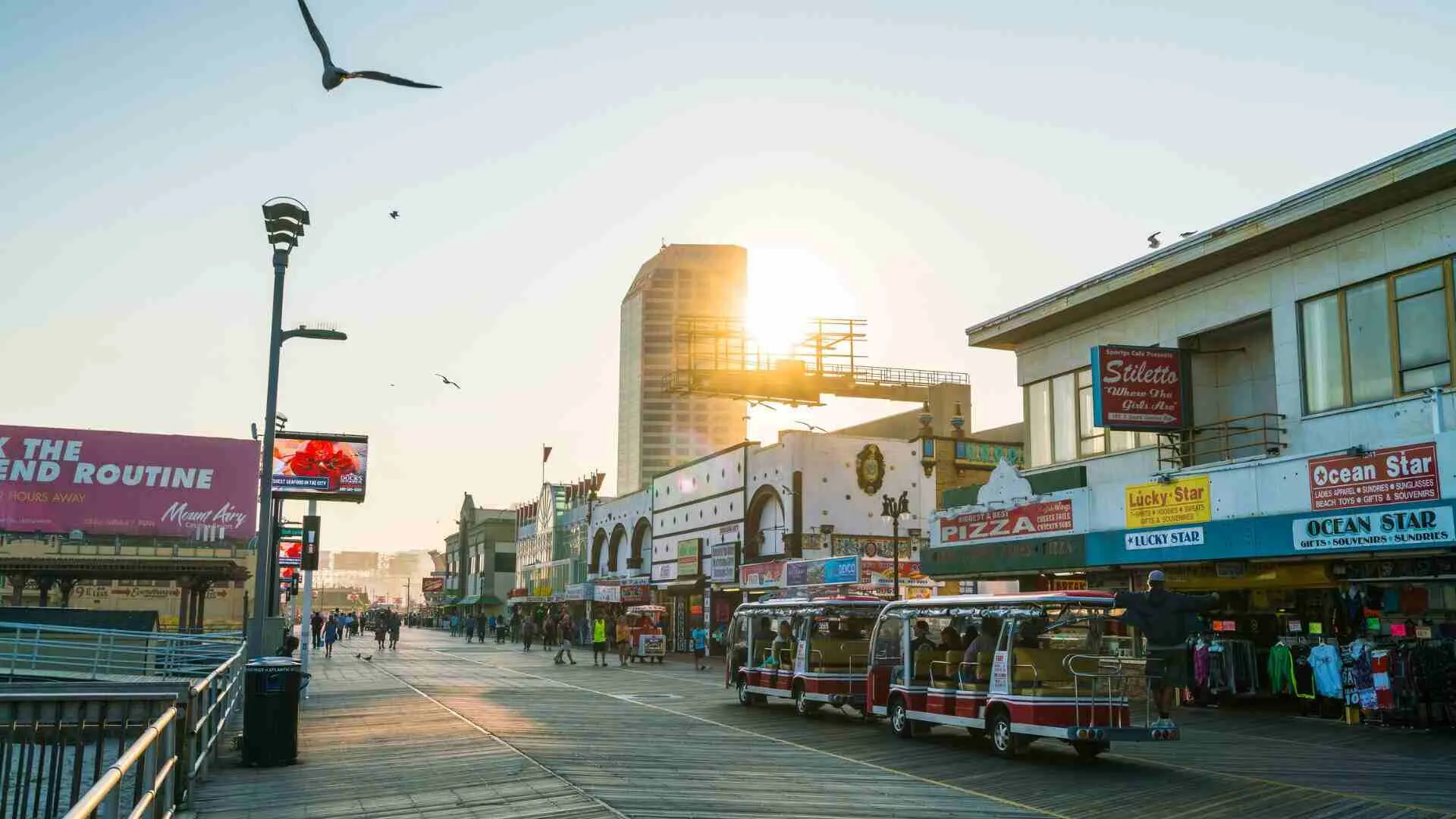 A scenic view of the boardwalk in Hasbrouck Heights, NJ, with shops, people, and rickshaw-like vehicles during sunset.