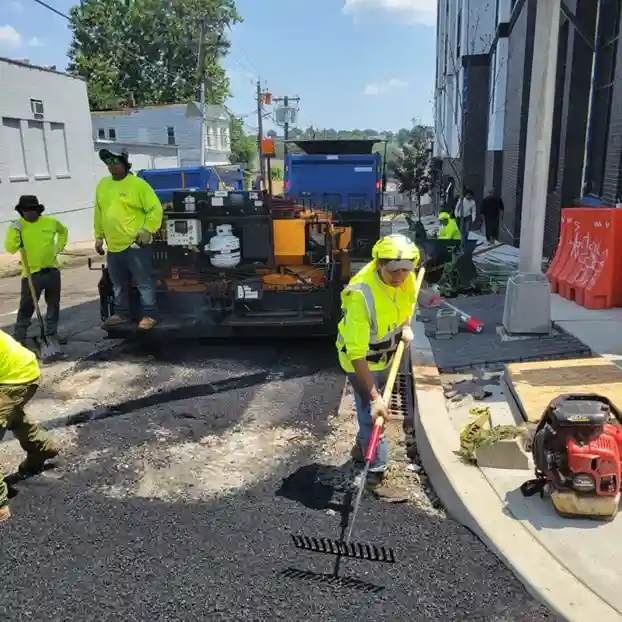 A worker using a rake to smooth the asphalt, while others are assisting with the paving equipment in the background